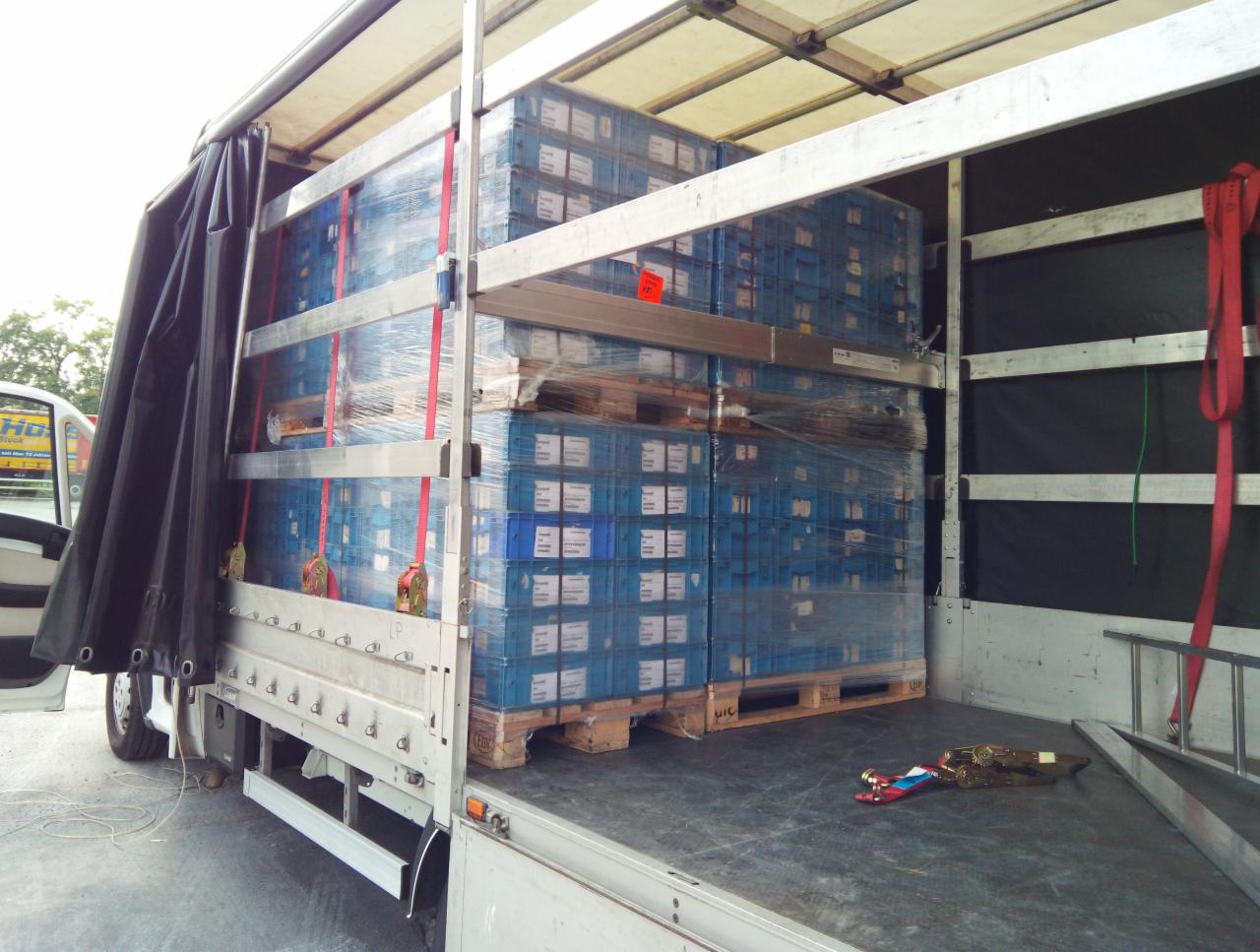 Wnętrze naczepy z załadowanymi paletami niebieskich skrzynek, zabezpieczonych folią i pasami, widok z tyłu, częściowo odsłonięta plandeka.