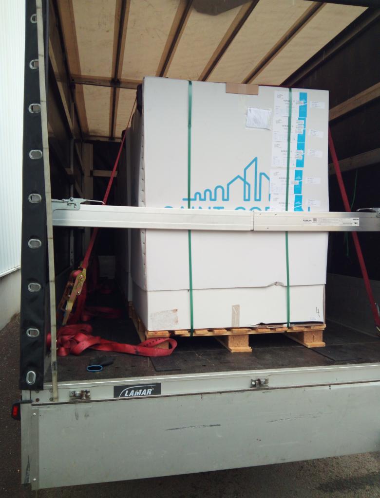 Wnętrze naczepy z ładunkiem zabezpieczonym czerwonymi pasami transportowymi, białe kartony na palecie z niebieskim logo firmy na górnym kartonie.