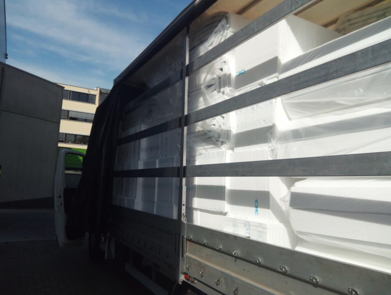 Ciężarówka z otwartą plandeką, wypełniona po brzegi białymi, spakowanymi elementami izolacyjnymi, widoczne budynki w tle.