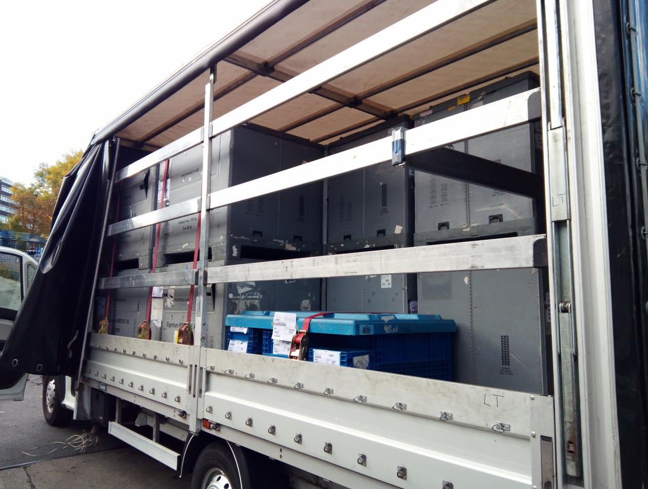 Wnętrze częściowo otwartej ciężarówki z ładunkiem zabezpieczonym pasami: szare metalowe skrzynie i niebieskie plastikowe pojemniki.