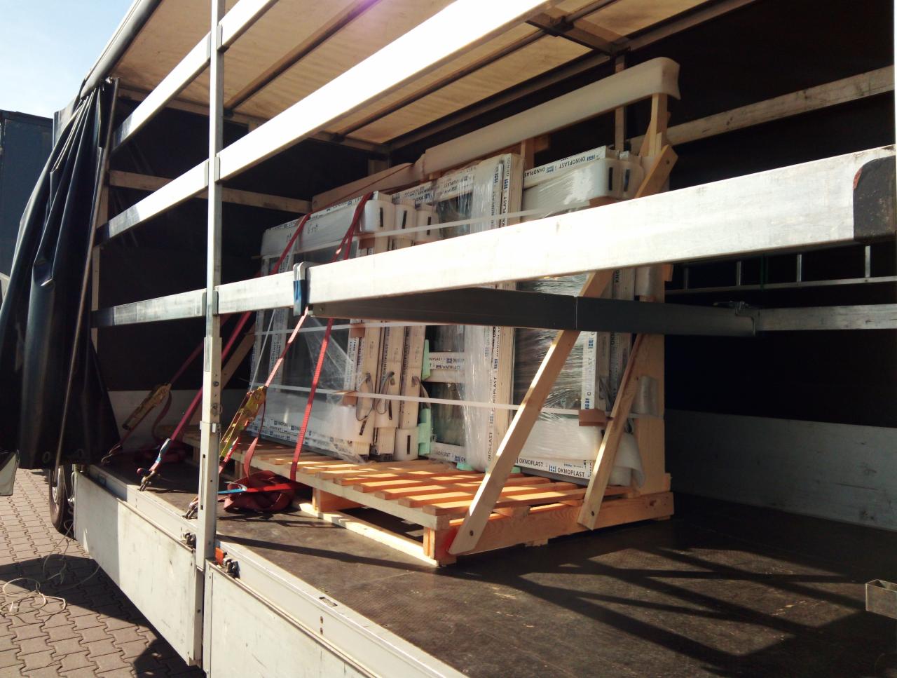 Okna PCV zabezpieczone folią i pasami transportowymi, umieszczone na drewnianych paletach wewnątrz naczepy ciężarówki z rozsuwaną plandeką.