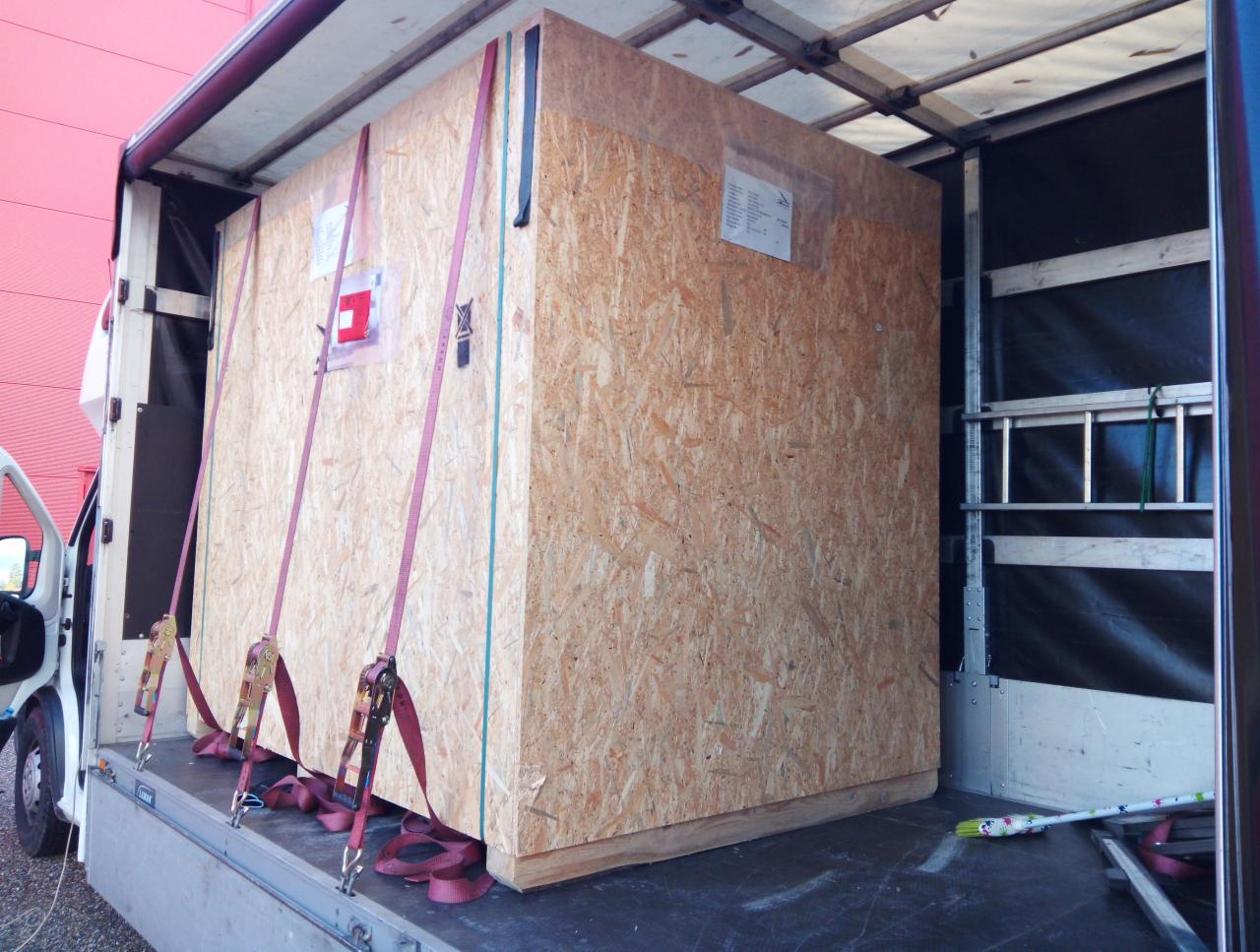 Duża, drewniana skrzynia zabezpieczona pasami wewnątrz przestrzeni ładunkowej samochodu dostawczego z otwartymi drzwiami.