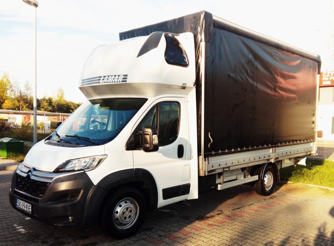 Biały bus dostawczy z czarną plandeką marki Lamar, zaparkowany na kostce brukowej w słoneczny dzień.