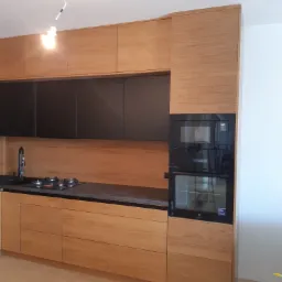 Kuchnia na wymiar w Krakowie z drewnianymi frontami szafek dolnych i górnych, czarne szafki wiszące, czarny blat i sprzęt AGD w zabudowie.