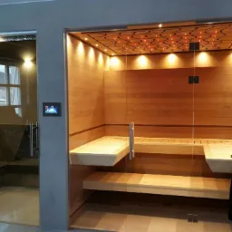 Nowoczesna sauna z drewnianym wnętrzem i oświetleniem sufitowym, obok przeszklona kabina prysznicowa z marmurowym wykończeniem.