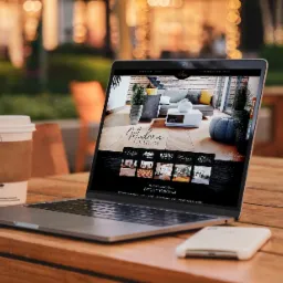 Laptop wyświetlający stronę internetową z kolekcją nowoczesnych podłóg na drewnianym stole na zewnątrz, obok kubek z kawą i smartfon.