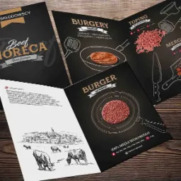 Rozłożone ulotki reklamowe produktów mięsnych, prezentujące burgery i merguez, z elementami graficznymi i informacjami o firmie Skłodowscy, na drewnianym tle.