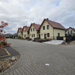 Nowe osiedle domów jednorodzinnych z beżową elewacją i czerwoną dachówką, widok z brukowanej drogi, na której leży biała kartka papieru. W tle pochmurne niebo.