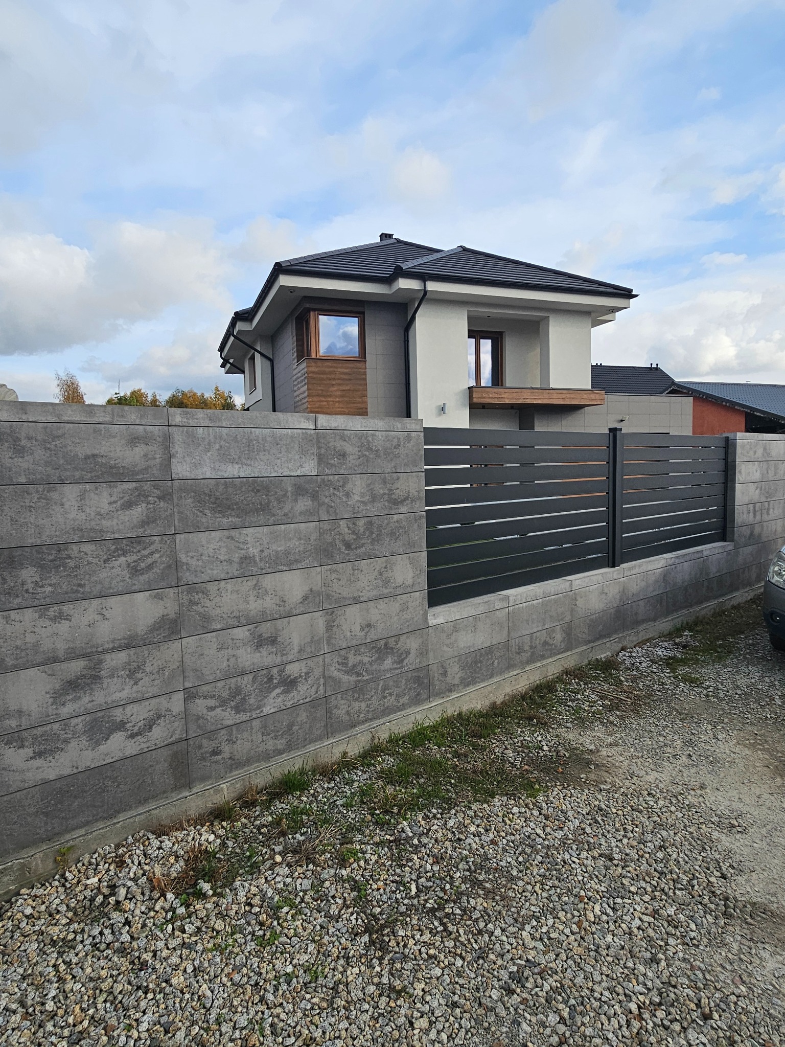 Nowoczesny dom jednorodzinny z szarą elewacją i grafitową stolarką okienną, otoczony ogrodzeniem z betonowych bloczków i metalowych paneli, widok z poziomu gruntu na żwirowym podłożu.