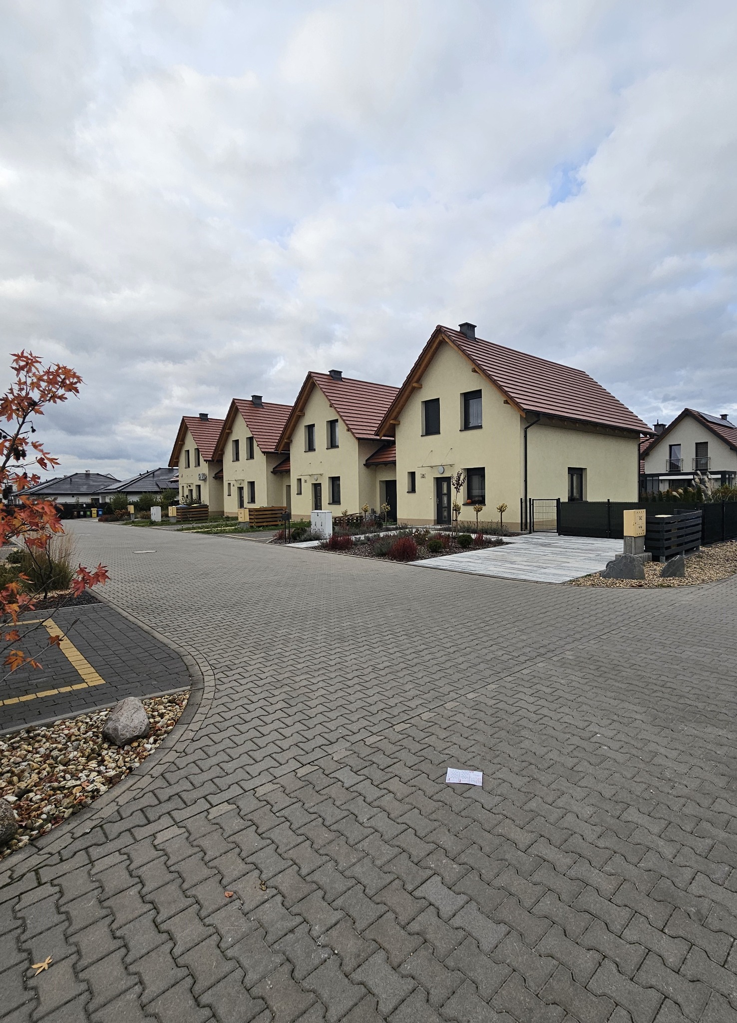 Nowe osiedle domów jednorodzinnych z beżową elewacją i czerwoną dachówką, widok z brukowanej drogi, na której leży biała kartka papieru. W tle pochmurne niebo.