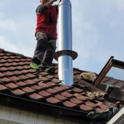 Instalacja błyszczącego, metalowego komina na dachu z czerwonej dachówki; pracownik w czerwonej kamizelce i zielonych butach uśmiecha się, stojąc na dachu.