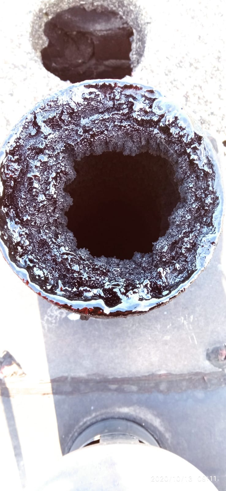 Zamarznięty wylot komina z widocznymi kryształkami lodu na wewnętrznej krawędzi, sfotografowany z góry w słoneczny dzień.