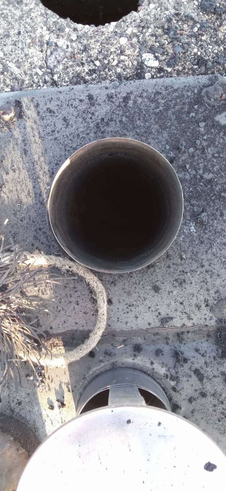 Wnętrze metalowego komina z widocznymi osadami sadzy, widok z góry na tle betonowego dachu z fragmentami luźnego gruzu i szczotką kominiarską obok.
