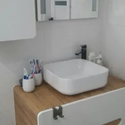 Nowoczesna łazienka z kwadratową umywalką nablatową, czarną baterią i szafką z drewnianym blatem oraz białymi frontami z subtelnymi uchwytami.