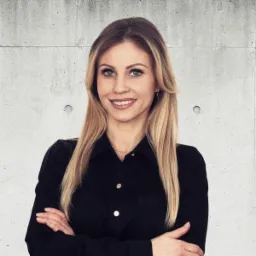 Portret uśmiechniętej blondynki w czarnej koszuli, ręce skrzyżowane, na tle surowej betonowej ściany.