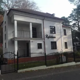 Dwupiętrowy budynek z balkonem i napisem 'Rybitwa' na ścianie, otoczony czarnym, ozdobnym ogrodzeniem, z widocznymi drzewami w tle.