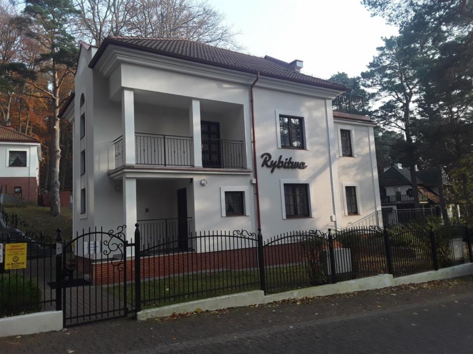 Dwupiętrowy budynek z balkonem i napisem 'Rybitwa' na ścianie, otoczony czarnym, ozdobnym ogrodzeniem, z widocznymi drzewami w tle.