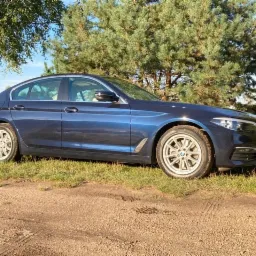 Niebieski sedan BMW serii 5 zaparkowany na trawiastym poboczu polnej drogi, widok z boku, z widocznym logo na feldze.