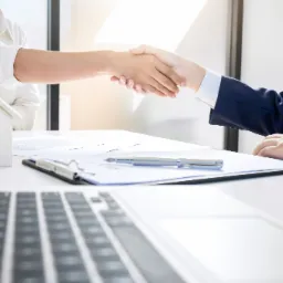Uścisk dłoni nad dokumentami i modelem domu, laptop na pierwszym planie, symbolizujący zawarcie umowy.