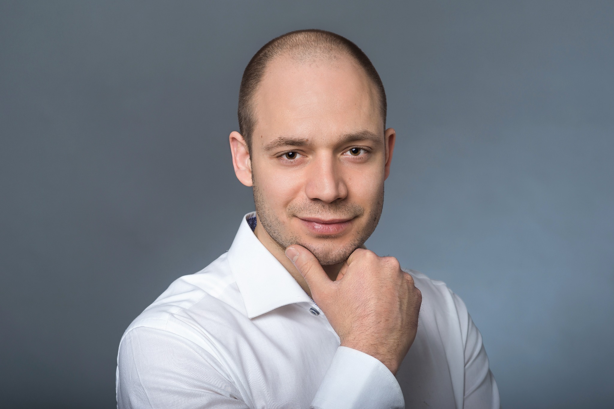 Portret mężczyzny w białej koszuli, z dłonią pod brodą, na szarym tle.
