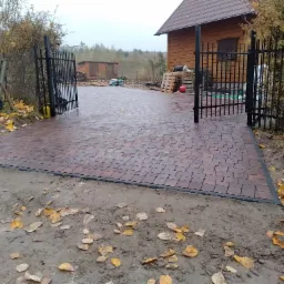 Nowo ułożona, brązowa kostka brukowa prowadząca do otwartej, czarnej bramy wjazdowej. Widoczne liście na ziemi i drewniany dom w tle.