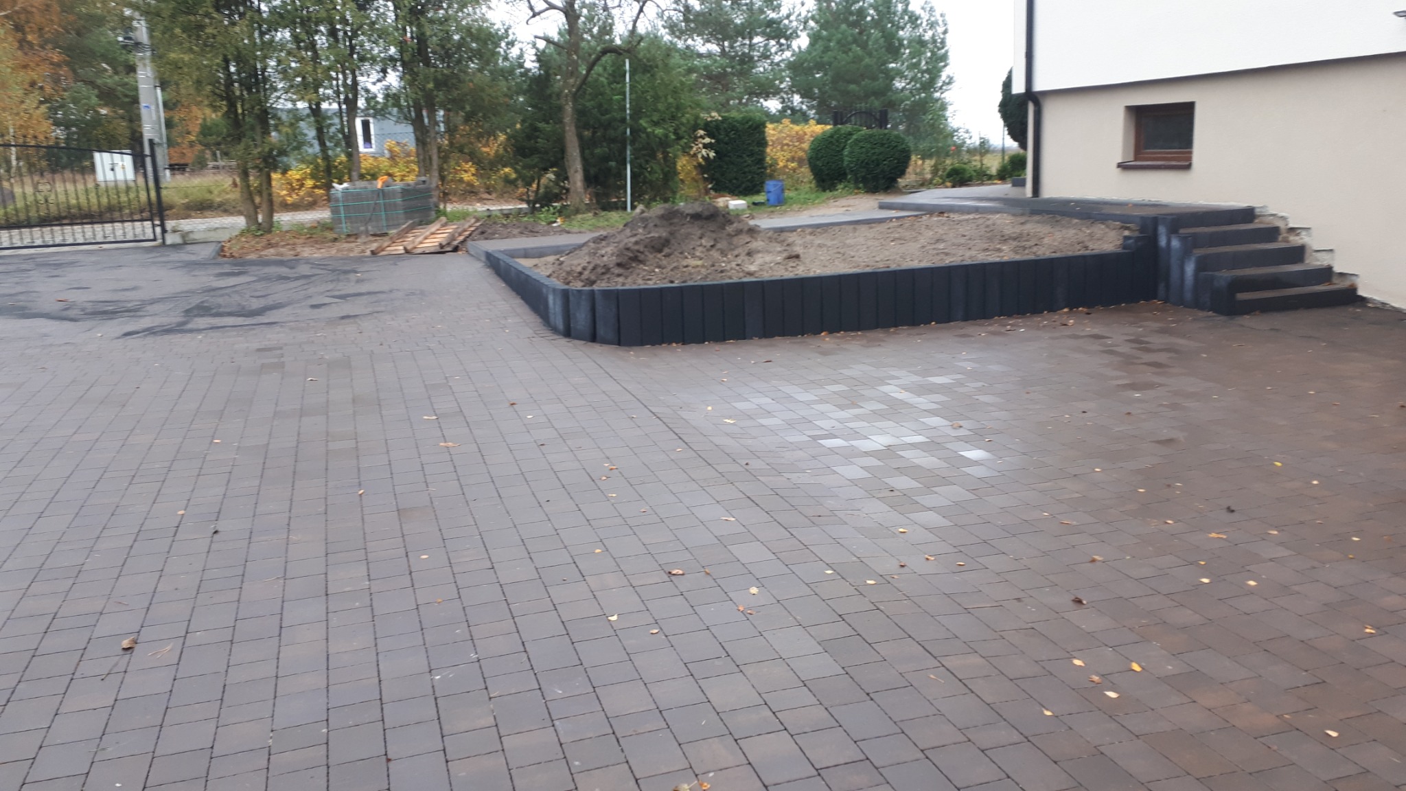 Ułożona kostka brukowa na podjeździe, z widocznym obrzeżem rabaty kwiatowej z ciemnych betonowych elementów oraz schodami wejściowymi do domu po prawej stronie. Na rabacie usypana ziemia.