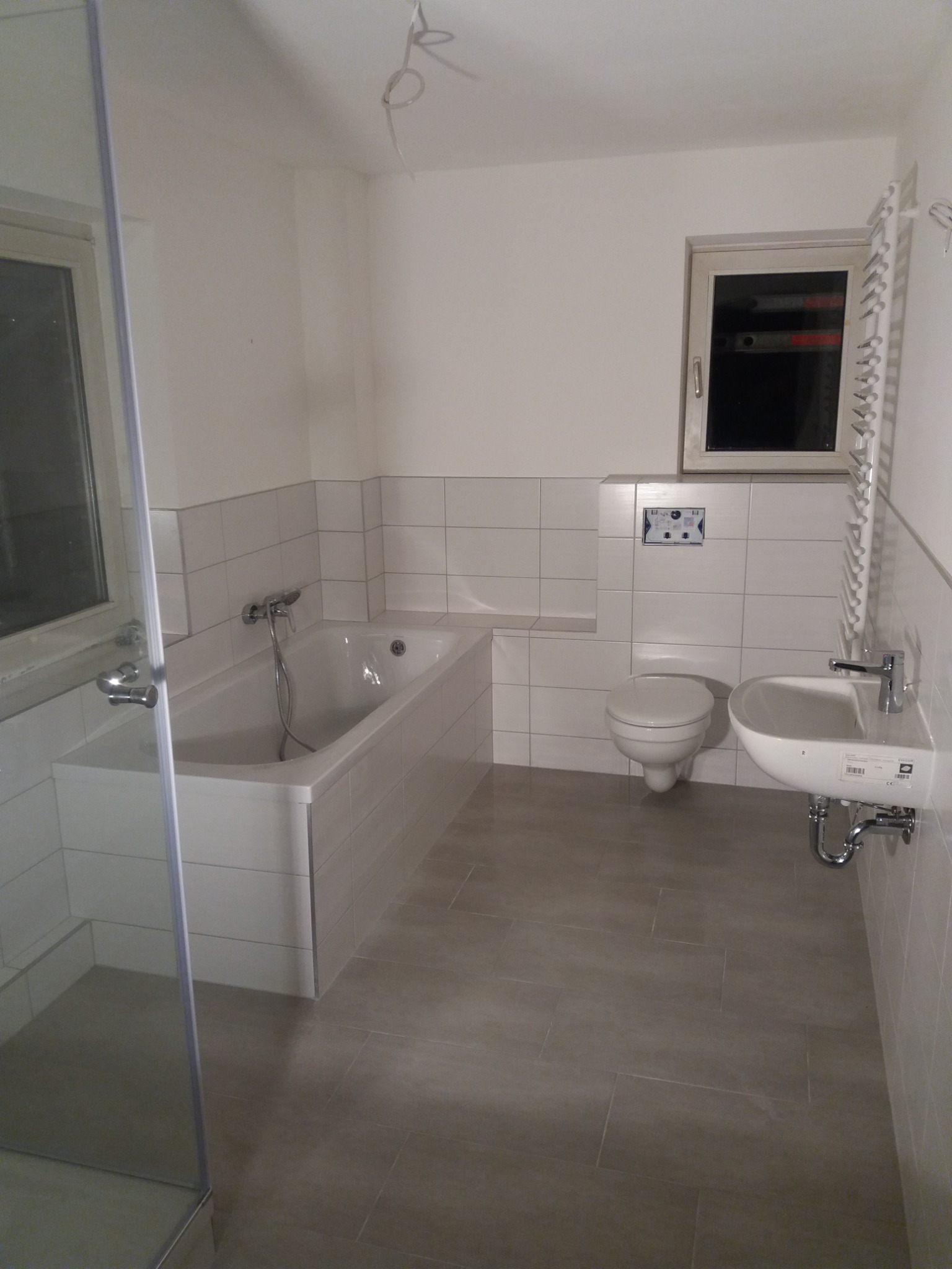 Jasna łazienka w trakcie wykańczania, widoczna wanna, umywalka, toaleta, grzejnik drabinkowy oraz szare płytki podłogowe i białe ścienne. Widoczne niedokończone podłączenia elektryczne na suficie.