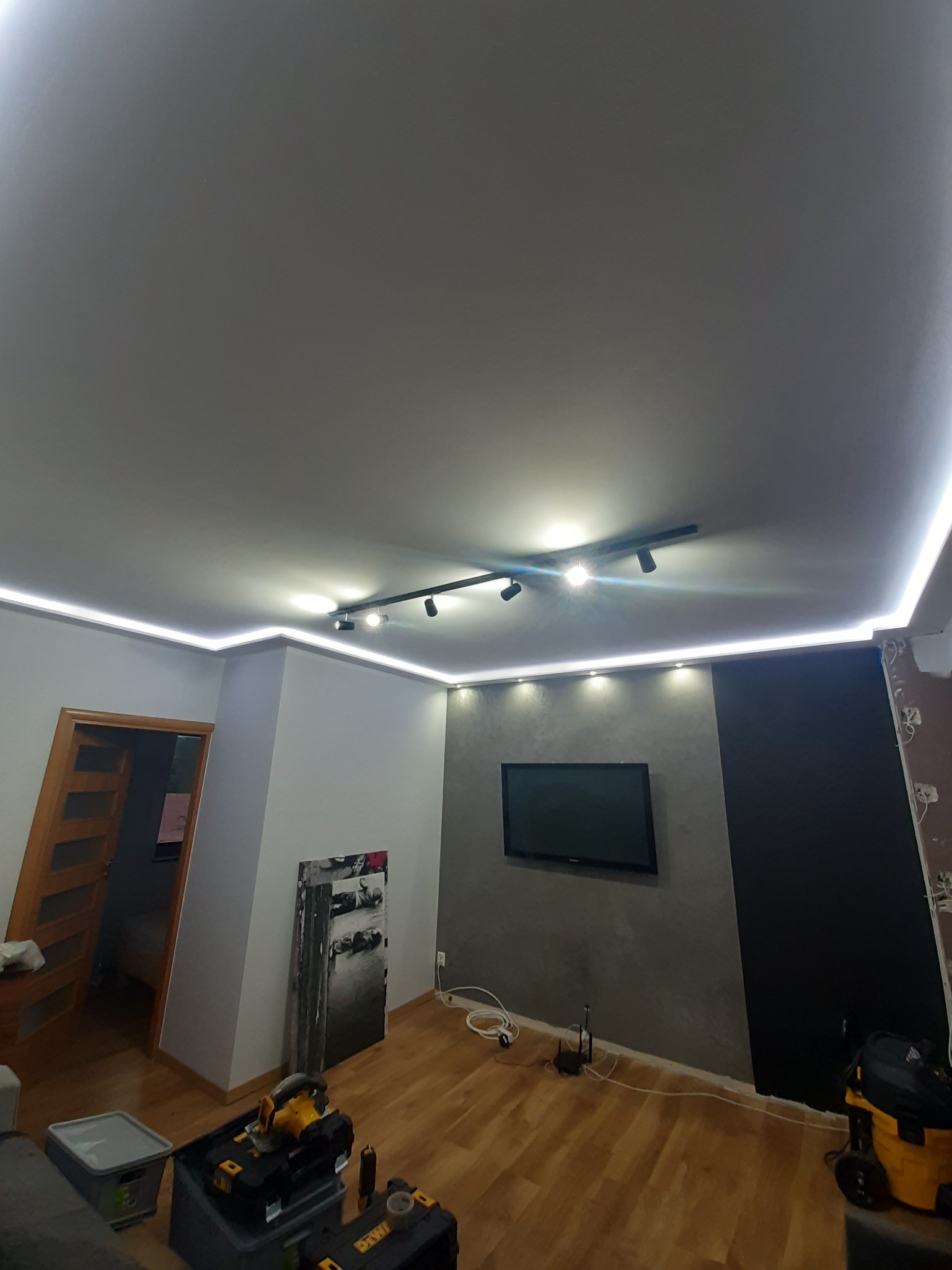 Pokój w trakcie remontu z widocznym oświetleniem LED na suficie, surową ścianą pomalowaną częściowo na szaro i czarno, narzędziami budowlanymi na podłodze i telewizorem na ścianie.