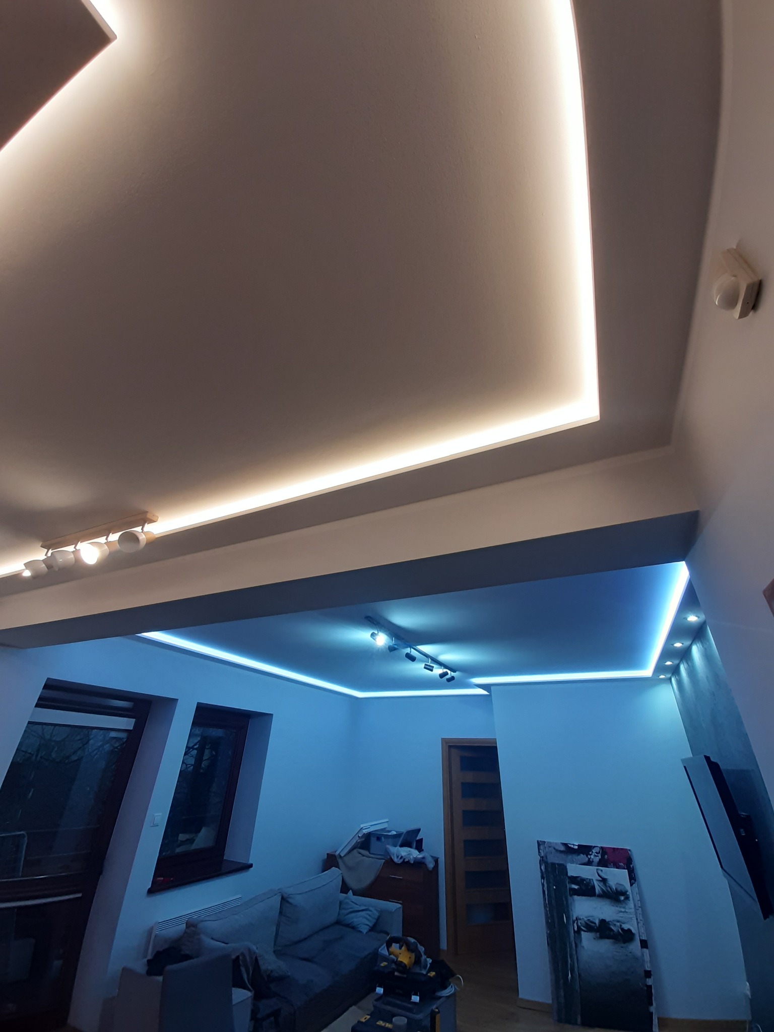 Dwupoziomowy sufit z oświetleniem LED: białe światło w górnej części i niebieskie w dolnej, widoczne lampy punktowe i fragment pokoju z meblami.