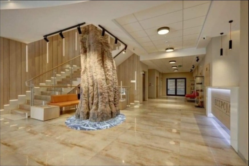 Recepcja hotelowa z dekoracyjnym pniem drzewa obłożonym kamieniem u podstawy schodów, oświetlenie punktowe, podłoga z płytek imitujących marmur.