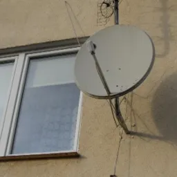 Biała antena satelitarna zamontowana na elewacji budynku obok okna, rzucająca cień na ścianę.