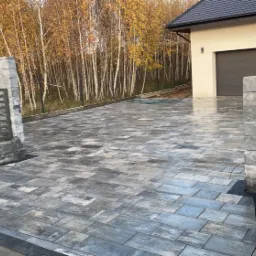 Ułożona kostka brukowa w Gdyni, prowadząca do domu z garażem, widoczna brama z szarymi słupkami i metalowymi przęsłami oraz drzewa w tle.