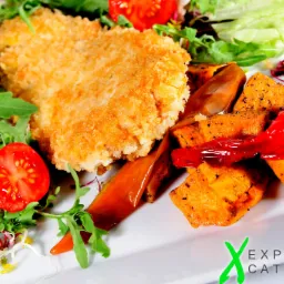 Elegancko podane danie: panierowany kotlet z sałatką z rukoli, pomidora i kiełków oraz pieczone bataty z papryką na białym talerzu. W prawym dolnym rogu widoczne logo firmy cateringowej.