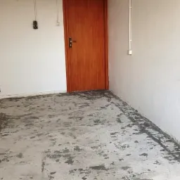 Przygotowanie podłoża pod wylewkę w pomieszczeniu z białą ścianą, brązowymi drzwiami i czarnym panelem sterowania.