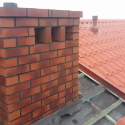 Nowo wybudowany komin z cegły klinkierowej z otworami wentylacyjnymi, stojący na dachu pokrytym blachodachówką w odcieniach czerwieni, widoczne elementy konstrukcji dachu.