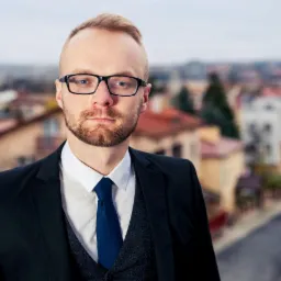 Portret mężczyzny w garniturze, okularach i z brodą na tle rozmytego krajobrazu miejskiego.