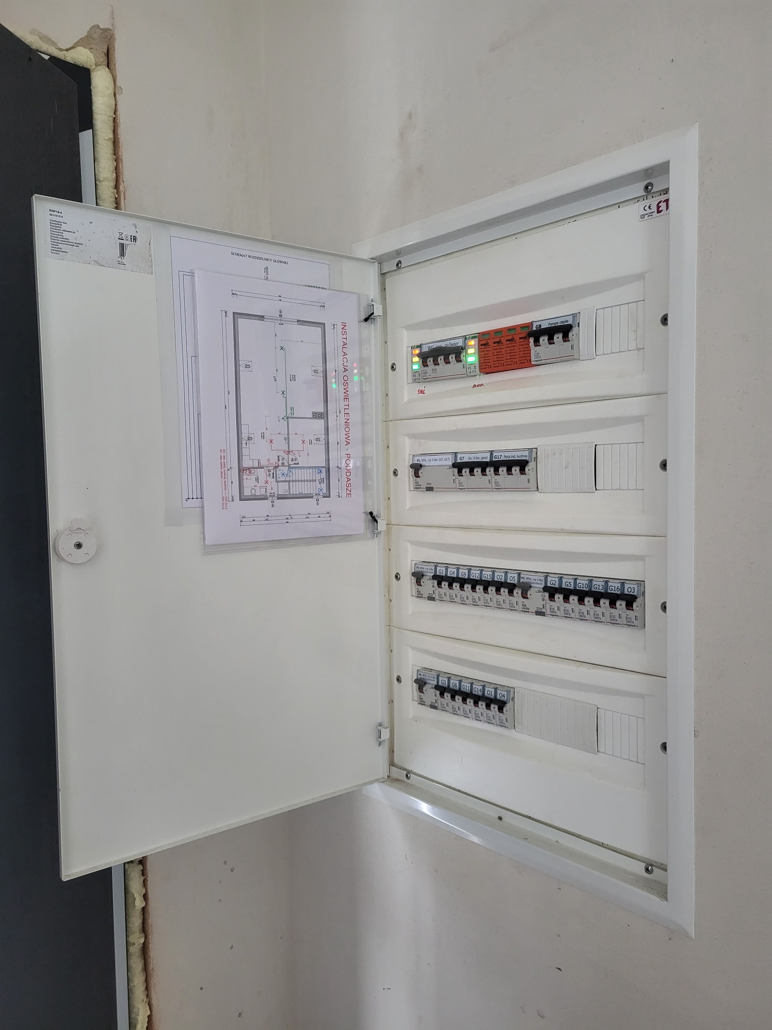 Otwarta skrzynka rozdzielcza z zamontowanymi bezpiecznikami i wyłącznikami, schemat instalacji oświetleniowej poddasza przyczepiony do drzwiczek.