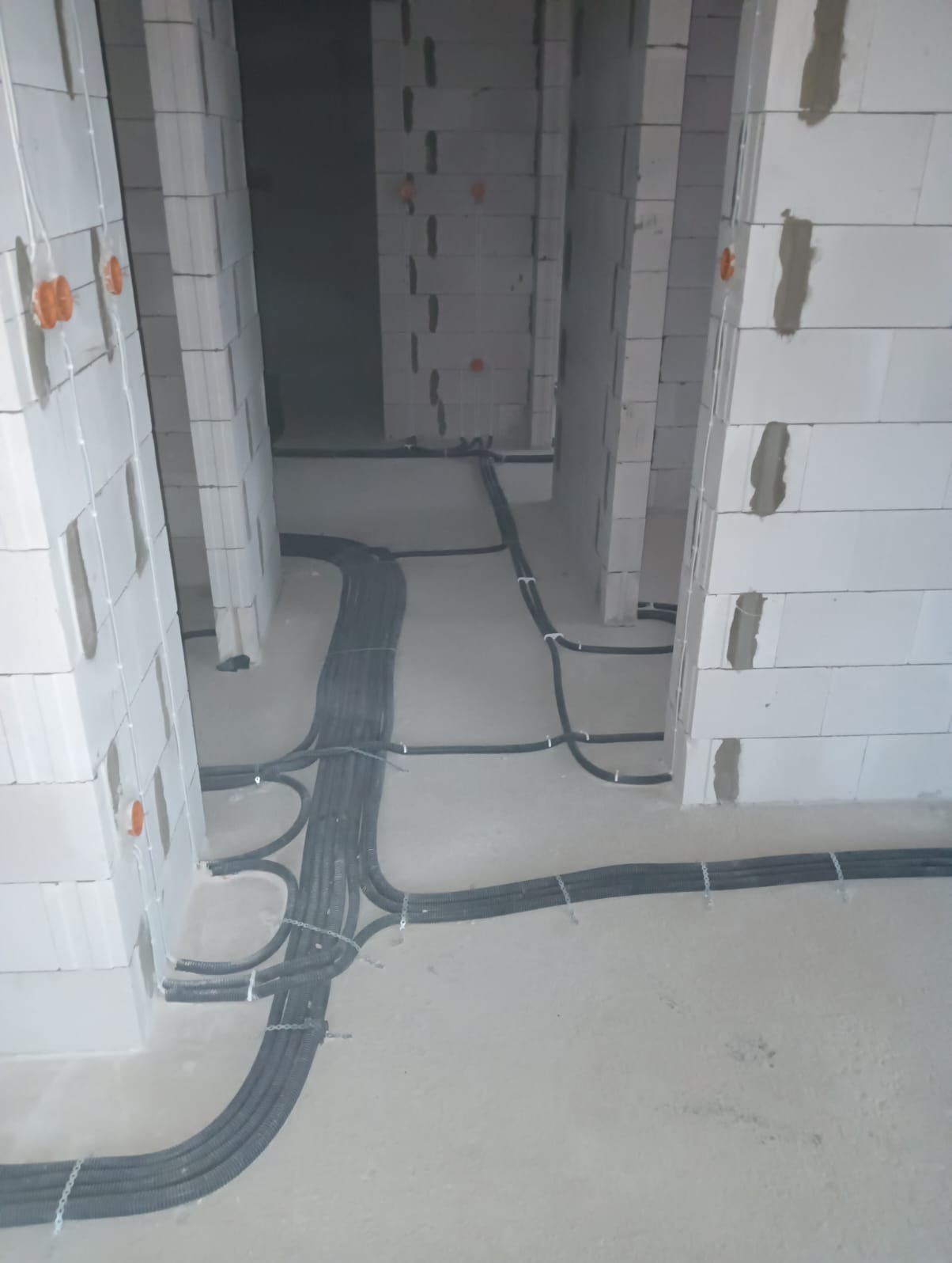 Wnętrze w trakcie instalacji elektrycznej w surowym domu z białego bloczka, widoczne peszle z okablowaniem ułożone na podłodze i puszki instalacyjne w ścianach.