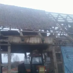 Częściowo rozebrany budynek gospodarczy z widoczną konstrukcją więźby dachowej i traktorem w tle, zabezpieczony plandeką.