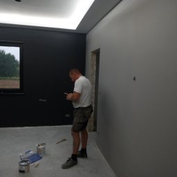 PROFESJONALNE WYKOŃCZENIA WNĘTRZ- PAWEŁ KOWAL - Mężczyzna w krótkich spodenkach i białej koszulce sprawdza telefon w trakcie malowania pokoju na dwa kolory: szary i czarny, widoczne puszki z farbą i pędzel.