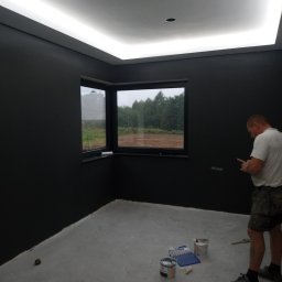 PROFESJONALNE WYKOŃCZENIA WNĘTRZ- PAWEŁ KOWAL - Pokój po malowaniu na ciemny kolor, widoczne puszki z farbą i pracownik sprawdzający coś na telefonie, oświetlenie sufitowe LED.