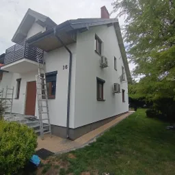 Świeżo pomalowana, biała elewacja domu z numerem 16, widoczne drabiny, okna z brązowymi ramami i roletami, klimatyzatory na ścianie, trawnik i zieleń w tle.