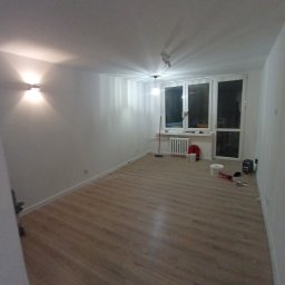 PROFESJONALNE WYKOŃCZENIA WNĘTRZ- PAWEŁ KOWAL - Pusty pokój po odświeżeniu, z panelami podłogowymi, pomalowanymi ścianami na biało, widoczne narzędzia malarskie i sprzęt grzewczy pod oknem.