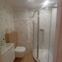 PROFESJONALNE WYKOŃCZENIA WNĘTRZ- PAWEŁ KOWAL - Nowoczesna łazienka z kabiną prysznicową z zaokrąglonymi drzwiami, umywalką z szafką, podwieszaną toaletą i płytkami imitującymi marmur na ścianach oraz drewnopodobnymi na podłodze.