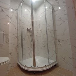 PROFESJONALNE WYKOŃCZENIA WNĘTRZ- PAWEŁ KOWAL - Szklana kabina prysznicowa z chromowanymi profilami w rogu łazienki, wykończonej białymi płytkami imitującymi marmur, widoczny fragment toalety.