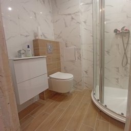 PROFESJONALNE WYKOŃCZENIA WNĘTRZ- PAWEŁ KOWAL - Nowoczesna łazienka z białymi marmurkowymi płytkami ściennymi, drewnopodobną podłogą, kabiną prysznicową z zaokrąglonym narożnikiem, wiszącą toaletą i szafką umywalkową z białą umywalką i armaturą.
