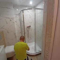 PROFESJONALNE WYKOŃCZENIA WNĘTRZ- PAWEŁ KOWAL - Montaż kabiny prysznicowej narożnej z chromowanymi profilami w łazience wyłożonej białymi płytkami imitującymi marmur, widoczny pracownik w jaskrawej koszulce, fragment niezabezpieczonej futryny po...