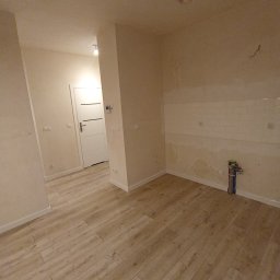 PROFESJONALNE WYKOŃCZENIA WNĘTRZ- PAWEŁ KOWAL - Wnętrze mieszkania w trakcie remontu: ułożone panele podłogowe, częściowo położone białe płytki w kuchni, ściany przygotowane do malowania, widoczne rury instalacji wodnej.