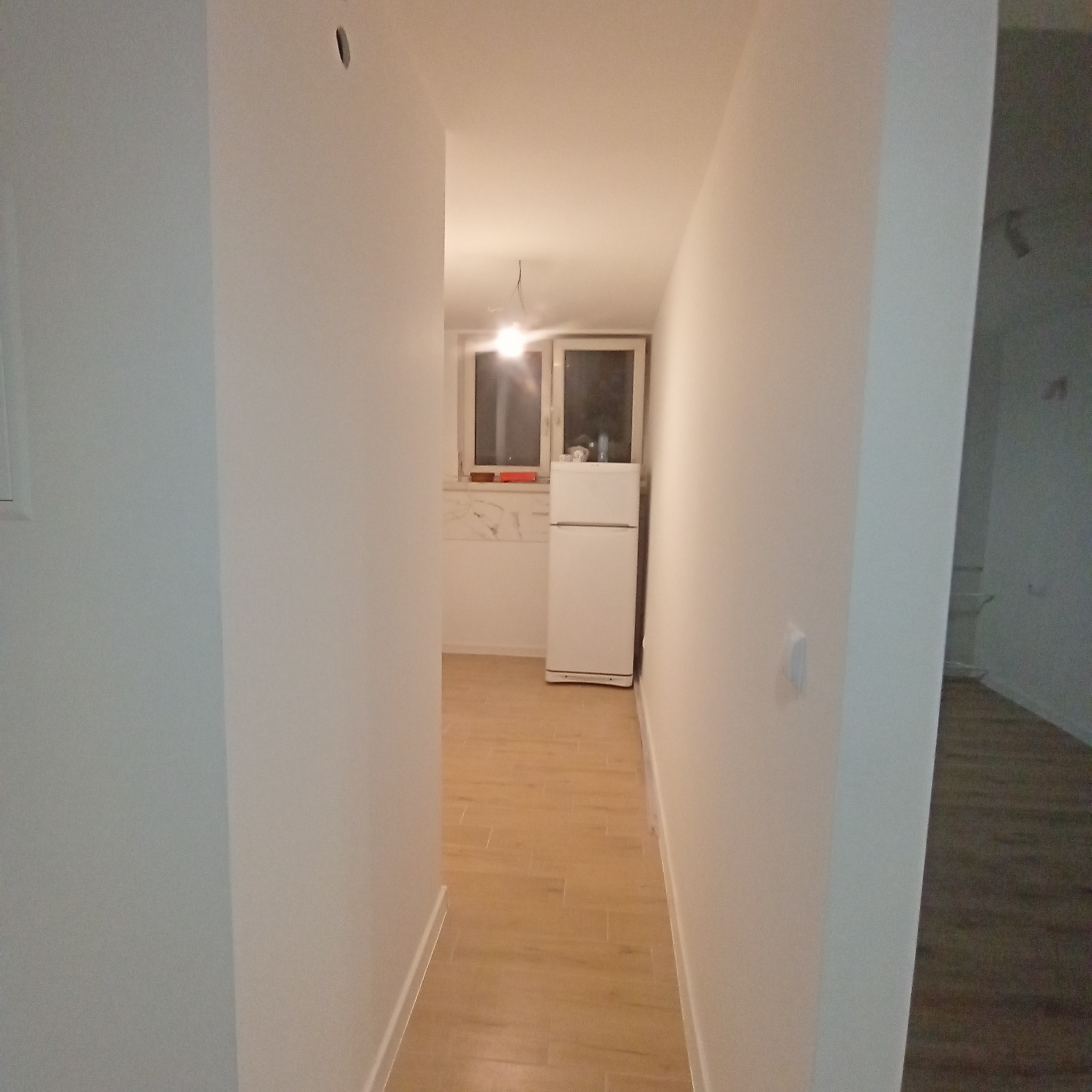 Wnętrze po odświeżeniu: widok z korytarza na jasną kuchnię z białymi ścianami, drewnopodobną podłogą i białą lodówką pod oknem.