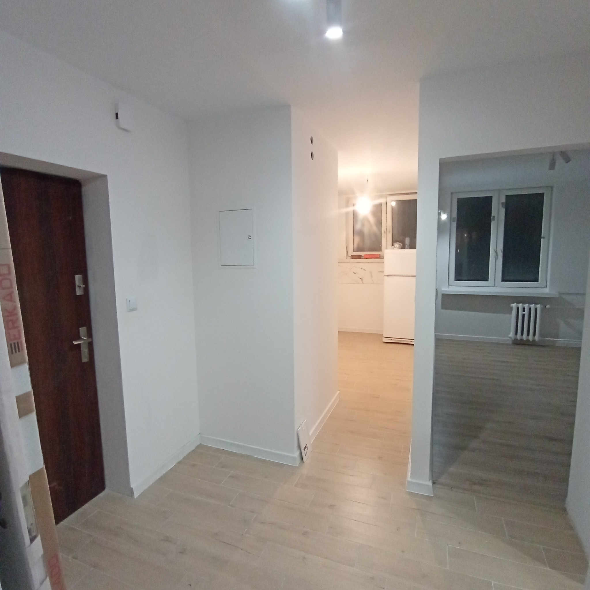 Jasne wnętrze po remoncie: białe ściany, drewnopodobne płytki podłogowe, widoczna kuchnia z białymi meblami i okno z grzejnikiem w drugim pokoju.